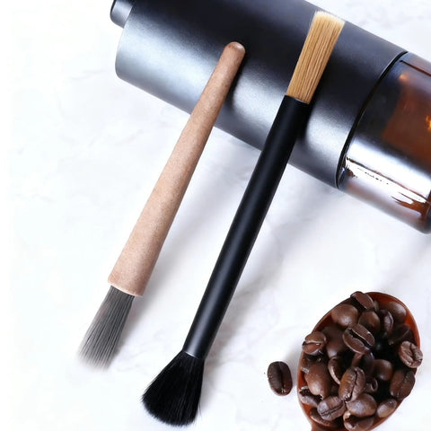 Two brushes from the Dual Head Coffee Grinder And Espresso Machine Cleaning Brush With Stiff Bristles For Deep Ground Residue And Grinder Gunk And Soft Bristles For Gentle Dusting On Surfaces set shown alongside a coffee grinder and beans.