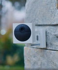 Battery Powered Wire Free Outdoor Security Camera Kit With Base Station 1080P Picture Night Vision Two Way Audio Works With Alexa And Google Assistant Easy DIY Install White With Camo Skin mounted on stone wall.