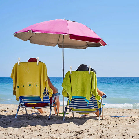 12 Pack Heavy Duty Plastic Clothes Pins Featuring Anti Rust Springs And Non Slip Silicone Pads holding towels on beach chairs under an umbrella.