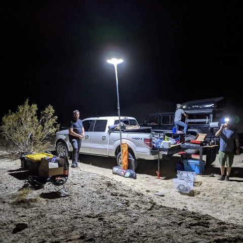 Beastly Bright 25000 Lumen LED Telescoping Camping Light illuminating a campsite with cars, supplies, and people at night.