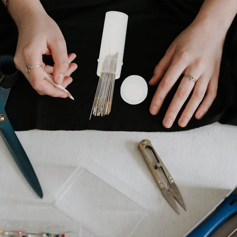 Two Pack Of Killer Heavy Duty Crewel Hand Sewing Needles With Super Easy Self Threading Eyes And A Hella Handy Storage Box on table with scissors.