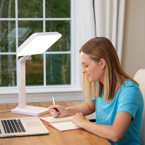 Woman using 10000 Lux Therapy Lamp For Mood And Energy Boost at desk, ideal for sunlight deprivation at home or office, white.