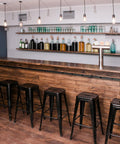 Modern bar area with wooden counter, high stools, and hanging lights, shelves stocked with bottles and glassware in the background.