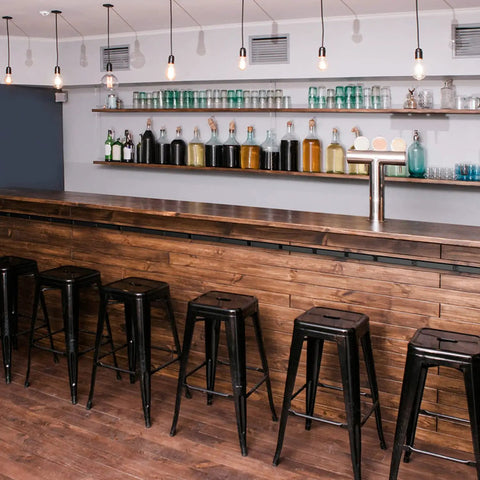 Modern bar area with wooden counter, high stools, and hanging lights, shelves stocked with bottles and glassware in the background.