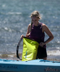 Person holding a Floating Waterproof Dry Bag Backpack 40L in bright yellow while standing next to a kayak on the shore.