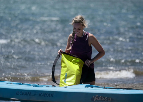 Person holding a Floating Waterproof Dry Bag Backpack 40L in bright yellow while standing next to a kayak on the shore.
