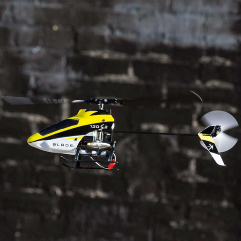 120S2 RC Helicopter BNF With SAFE Technology in flight, lightweight carbon fiber airframe, against a dark backdrop