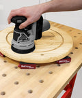 Triangle Bench Cookie Work Grippers 4 Pack Non Slip Project Supports For Woodworking And Painting Elevates Projects About 1.2 Inches On The Platform And 3 Inches With Built In Painting Supports For Edge Access And No Clamp Needed Setup showing a sander on wooden disc supported by red triangle grippers on a workbench.