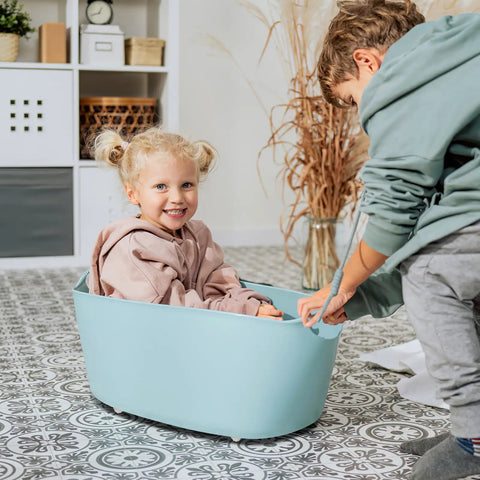 Child inside a blue plastic bin being pulled by another child, indoors on patterned flooring.