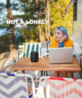 Person seated at outdoor table with a laptop and 15W Portable Bluetooth Speaker With Dual True Wireless Stereo For Party Vibe, Loud Crisp Highs And Deep Bass, IPX6 Waterproof Shower Speaker, Bluetooth 5.0, Dual Pairing For Home Theater And Gaming On The Go. Surrounded by trees and colorful chairs.