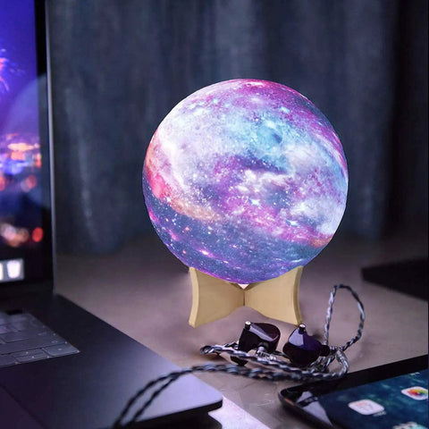 Galactic-themed LED night light globe on wooden stand next to a laptop, displaying colorful cosmic patterns.