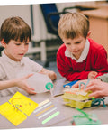 Two boys engaged with a yellow conductivity experiment kit, exploring electrical flow principles in a classroom.