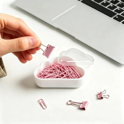 Hand holding a pink binder clip over a 12 Pack Cloud Paperclip Holder With Snug Lid Mini Plastic Desk Organizer with cloud design.
