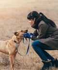 Person crouching in field with a dog on a leash, interacting closely, both wearing coats.