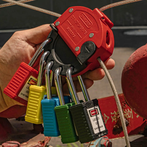 Hand holding multiple colored locks attached to a red lockout device in an industrial setting.