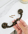 Hand holding Drop Bail Drawer Pull 5 Inch Center To Center 128 Mm Hole Spacing Includes 2 Screws Antique Brass Finish Floral Design