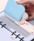 Hand using Three Hole Paper Puncher For Loose Paper In A4 A5 A6 A7 B5 Sizes, Handheld 5 Sheets Capacity on a notebook, showing alignment scale.