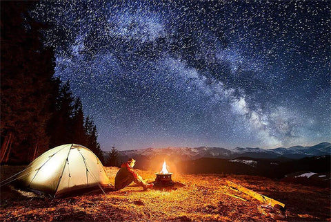 Man sitting by No Mess Smokeless Round Propane Fire Pit For Outdoors with tent under starry sky in mountains.