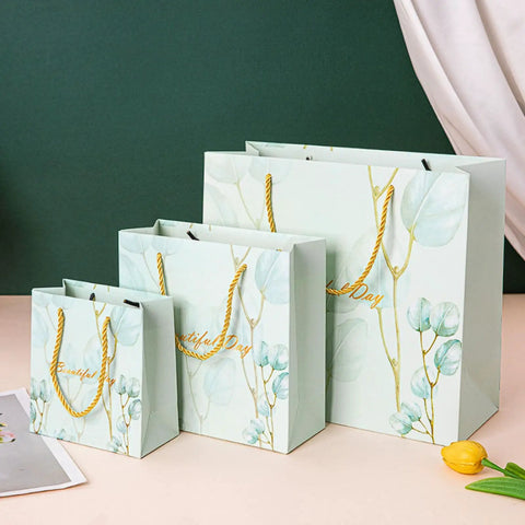Three floral paper bags, small to large, with yellow rope handles on a table, matching the Blue Paper Bags Set Of 30 Premium 3D Cut Totes For Shopping Clothing Jewelry And Souvenirs Includes 10 Small 10 Medium 10 Large Great For Grab And Go Gifts And DIY Packaging.