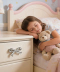 Pink And Silver Bow Dresser Knobs For Kids Rooms on drawer, girl asleep with teddy in nursery setting
