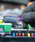 Close-up of a gloved hand holding a purple fuse near an electrical circuit board, part of a 100 Piece Car Fuse Assortment Kit With Tester And Puller Tool For Standard Mini Low Profile Blade Fuses Auto Truck Boat Electrical Replacement And Repair.