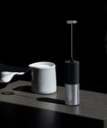 Mini Handheld Milk Frother Wand With Stainless Steel Whisk Power Packed 13,000 RPM Battery Powered Drink Mixer For Coffee Lattes Cappuccinos Matcha Hot Chocolate Iced Coffee And More Black No Stand Gift For Coffee Lovers, shown on a table beside a milk jug.