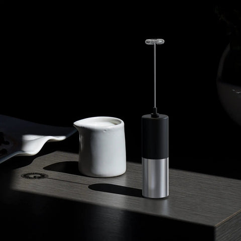 Mini Handheld Milk Frother Wand With Stainless Steel Whisk Power Packed 13,000 RPM Battery Powered Drink Mixer For Coffee Lattes Cappuccinos Matcha Hot Chocolate Iced Coffee And More Black No Stand Gift For Coffee Lovers, shown on a table beside a milk jug.