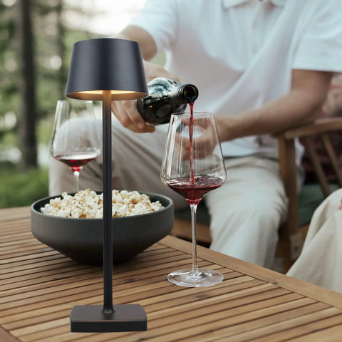 Must Have Rechargeable LED Desk Lamp Cordless With Touch Dimming 360 Degree Lighting Lightweight Aluminum Gold Finish Outdoor Ready Table Light for indoor use next to a bowl of popcorn and wine glasses on a wooden table.