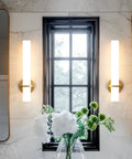 Two Pack Gold Bathroom Wall Sconces 18 Inch Modern LED Dimmable Wall Lights With Acrylic Lampshades Brushed Gold Finish For Bedroom Living Room Hallway Hardwired installed beside a window in a modern bathroom.