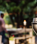 65 Inch Outdoor Torch With Amped Up Extra Large Flame For Patio Garden Lawn And Backyard Weather Resistant Refillable And Decorative Torch In Silver Finish Twist And Pour Refilling For Mess Free Fueling set against a blurred outdoor gathering.