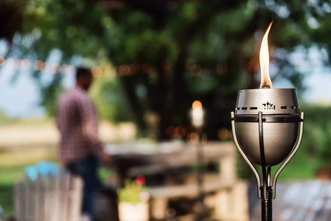 65 Inch Outdoor Torch With Amped Up Extra Large Flame For Patio Garden Lawn And Backyard Weather Resistant Refillable And Decorative Torch In Silver Finish Twist And Pour Refilling For Mess Free Fueling set against a blurred outdoor gathering.