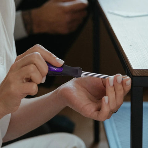 Person using a screwdriver from the 152 Piece Basic Household Hand Tool Set With Toolbox Storage Case All Purpose Home Tool Kit Includes Essential Tools For Office And College Repairs Purple on a table corner.
