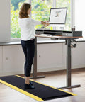 Anti Fatigue Standing Mat For Workshops 3 By 5 Feet Beefed Up Industrial Grade Ergonomic Floor Mat Nearly One Inch Thick With Non Slip Surface And Yellow Border For Leg And Back Pain Relief On Long Shifts in use at a standing desk.