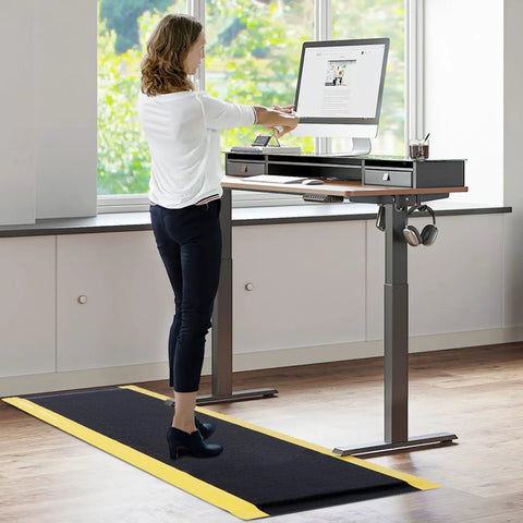 Anti Fatigue Standing Mat For Workshops 3 By 5 Feet Beefed Up Industrial Grade Ergonomic Floor Mat Nearly One Inch Thick With Non Slip Surface And Yellow Border For Leg And Back Pain Relief On Long Shifts in use at a standing desk.