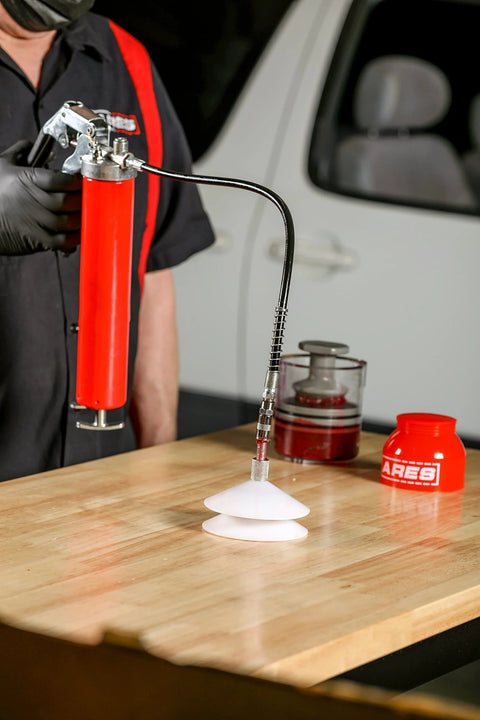 Side view of ARES bearing packer kit: red grease gun, hose, and two white cone-shaped packing cones on a wood table.
