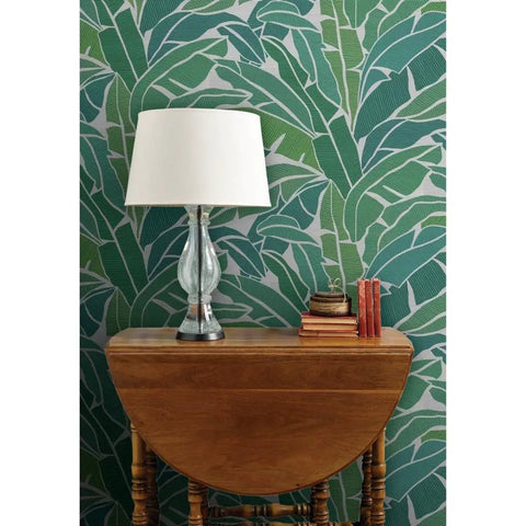 Top Shelf Tropical Leaf Wallpaper Exotic Plant Design Type II on a green wall behind a wooden table with a lamp and books.