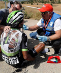 Front view of red first aid kit pouch opened on pavement with bandages beside a cyclist receiving care.