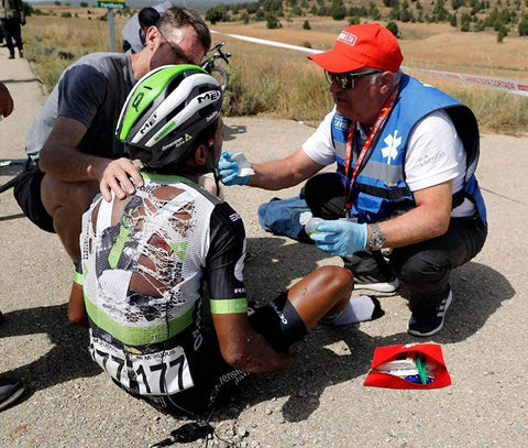 Front view of red first aid kit pouch opened on pavement with bandages beside a cyclist receiving care.