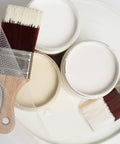 Open cans of Drop Cloth Chalk Mineral Paint For Furniture In Matte Linen White 16 Oz with paintbrush on white surface