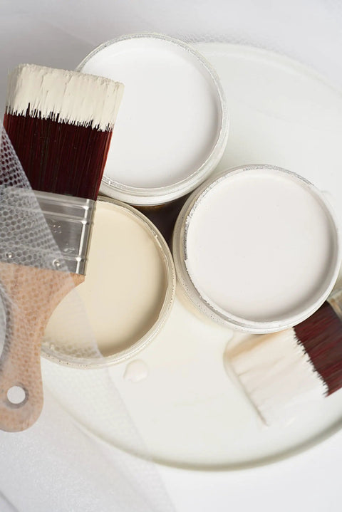 Open cans of Drop Cloth Chalk Mineral Paint For Furniture In Matte Linen White 16 Oz with paintbrush on white surface