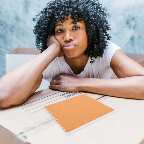 Person resting on a cardboard box, highlighting Heavy Duty Paper Packing Tape For Boxes With Easy Tear Strip And Writable Surface.