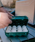 Hands securing Tough 12-Egg Plastic Carrier With Locking Lid, Molded Handle, And Secure Fit For Camping, Road Trips, And Meal Prep on table.