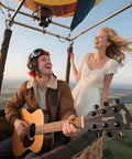 Two people smiling in a hot air balloon with a person playing a guitar labeled 'Gipson', open sky view.
