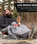 Man sitting outdoors on Low Profile Folding Camping Chair With Carry Bag Lightweight Breathable Mesh Seat For Camping Beach Picnic Barbecues And Sporting Events Easy Transport And Cushioned Armrests Black, wrapped in a blanket.