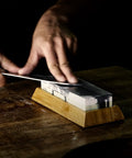 Using a Toishi Sharpening Whetstone With 1000 And 3000 Grit For Knife Edge Maintenance on a bamboo stand, dark background.