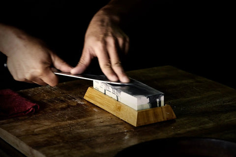 Using a Toishi Sharpening Whetstone With 1000 And 3000 Grit For Knife Edge Maintenance on a bamboo stand, dark background.
