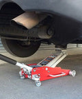 Red hydraulic floor jack with black handle lifting a car; exhaust pipe and wheel visible beneath.