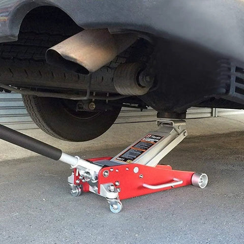 Red hydraulic floor jack with black handle lifting a car; exhaust pipe and wheel visible beneath.