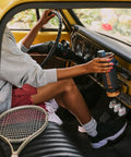 Person holding a Rock Solid Leakproof Shaker Bottle With Connected Hinge Cap And Fast Flow Spout in a vintage car interior.