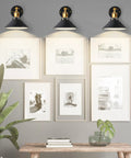 Three adjustable matte black wall sconces with gold stem illuminating a gallery hallway, showcasing modern decor with framed art.